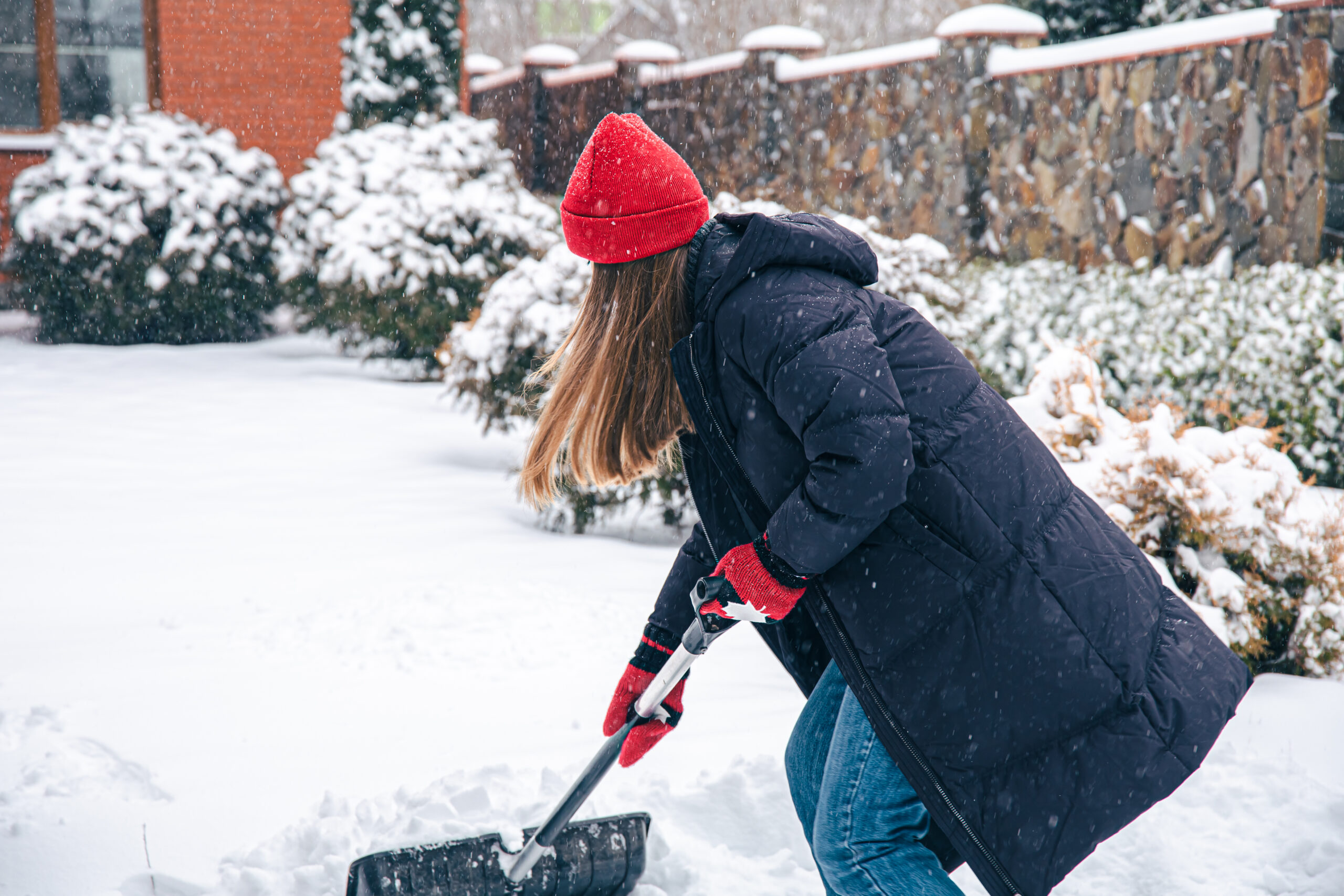 A young woman in a red hat, red mittens and a black jacket cleans snow in the yard in snowy weather, copy space.